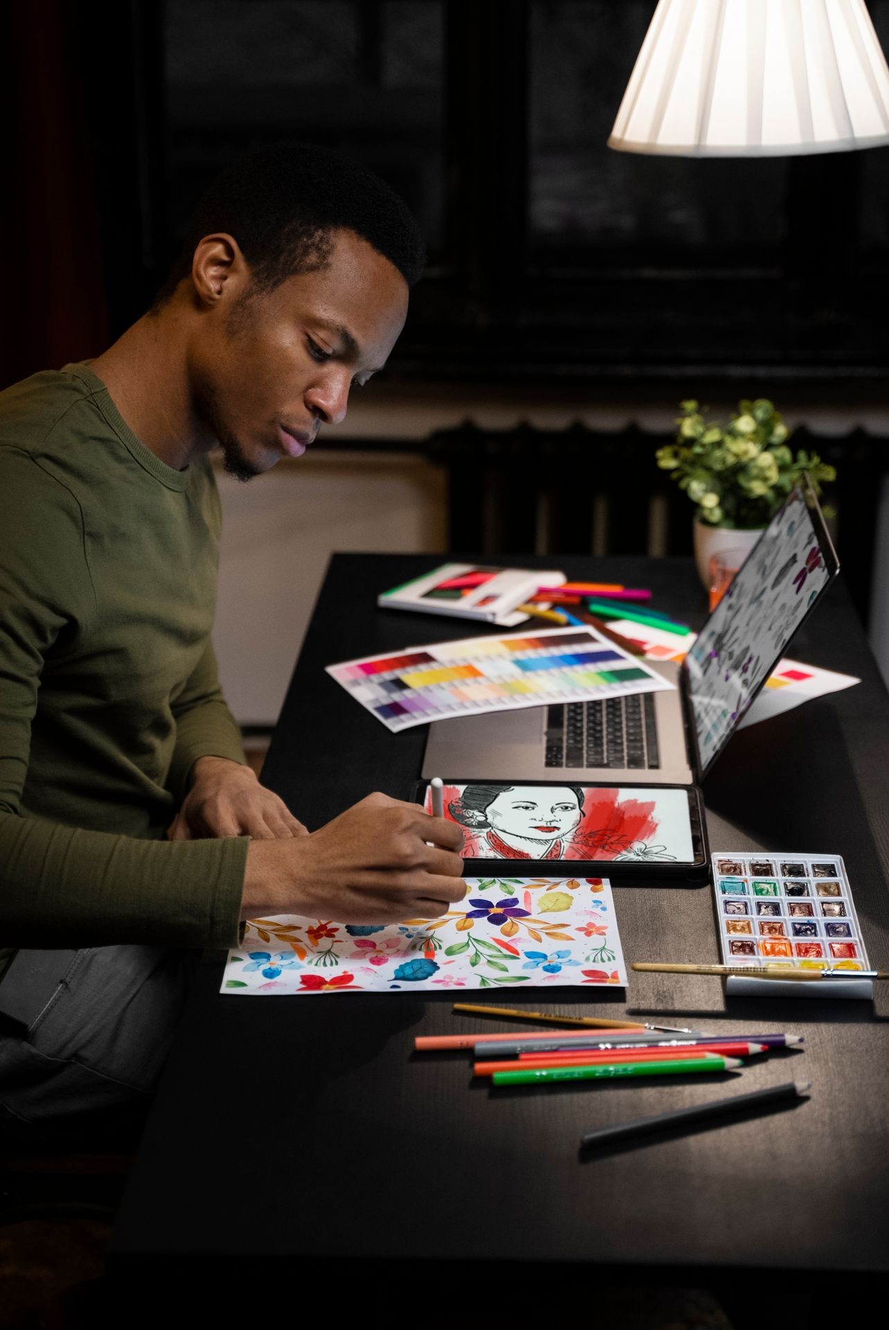 Artist drawing colorful flowers at a desk with laptop, tablet, and art supplies under a lamp.